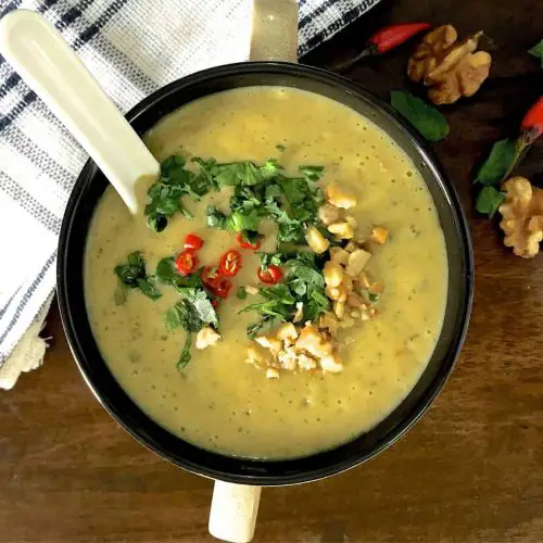 Overhead view of a cup of yellow curried chilli zucchini soup, garnished with slices of red chillies, coriander, parsley and mint leaves and walnuts. A white napkin striped with dark blue to the left, and walnuts and red chillies to the right