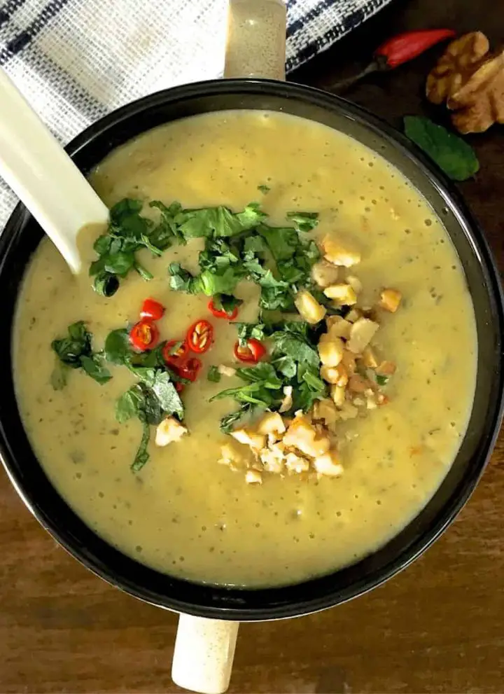 Overhead view of a cup of yellow curried chilli zucchini soup, garnished with slices of red chillies, coriander, parsley and mint leaves and walnuts. A white napkin striped with dark blue to the left, and walnuts and red chillies to the right