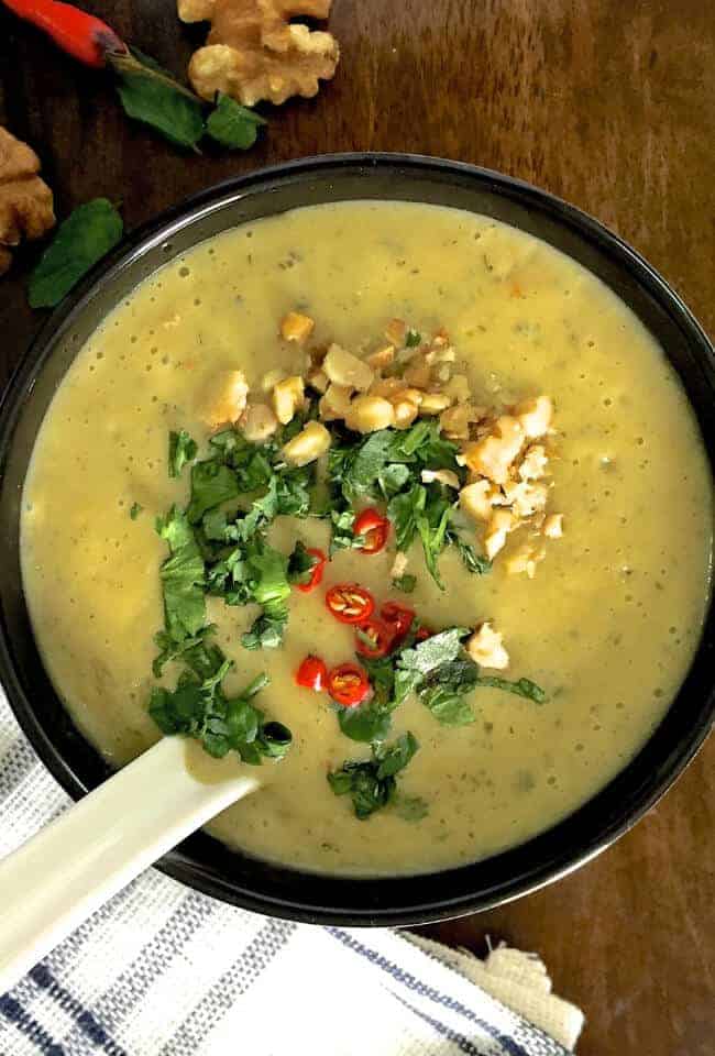 A cup of mustardy yellow Chilli Zucchini Soup with Walnuts and curried with chilli powder and Turmeric. Garnished with red chilli slices, walnuts and fresh herbs