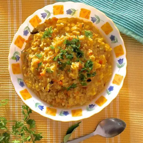 Bright yellow khicidi for the new mom, made from rice and lentils cooked together with vegetables, garnished with coriander leaves, on a white plate with a yellow and blue design at the edge. Coriander leavs and a steel spoon in the foreground. A blue green napkin on the top left and all set on ayellow mat