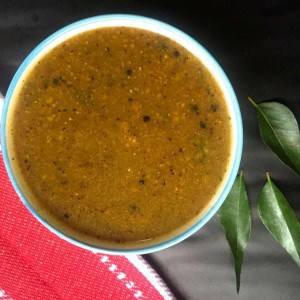 Pathiya Milagu Kuzhambu, a brown fluid gravy from pepper paste, in a blue bowl. A red napkin to the bottom left of the bowl and curry leaves scattered on the right.