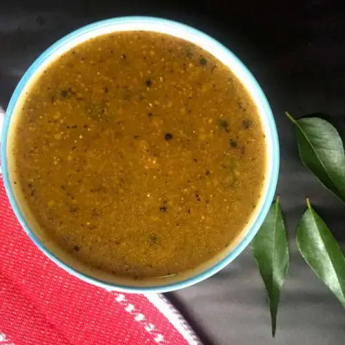 Pathiya Milagu Kuzhambu, a brown fluid gravy from pepper paste, in a blue bowl. A red napkin to the bottom left of the bowl and curry leaves scattered on the right.