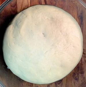 A round sphere of Basic Pizza Dough rising in a glass bowl on a brown wooden background