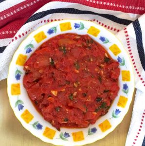 Tomato basil sauce in a white bowl with the edge patterned with purple flowers adn yellow squares, on a yellow background and with a red, black and white sriped napkin above