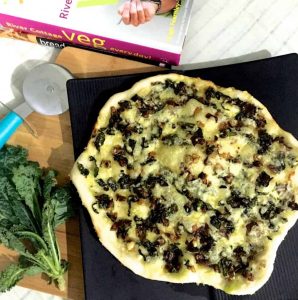 Easy homemade Kale and onion pizza, with the sauteed kale and onion and cheeze topping the pizza. A blue pizza cutter, A bunch of green kale and book with a pink binding seen around the pizza along with a white striped napkin