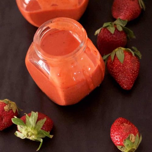 2 little glass jars of orangey pink strawberry poppyseed salad dressing, with strawberries strewn around on a black background