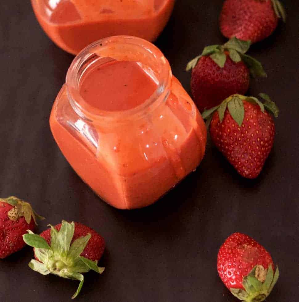 2 little glass jars of orangey pink strawberry poppyseed salad dressing, with strawberries strewn around on a black background