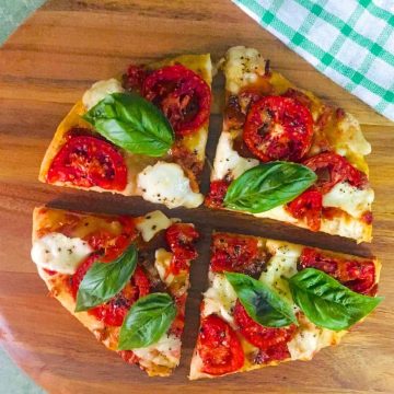 4 slices of a round Margherita Pizza/ Tomato Mozzarella Pizza with bright red tomato and fresh green basil leaves on a brown cutting board, with a green and white checked napkin to the side