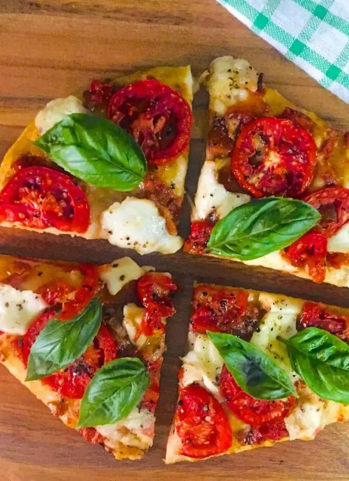 4 slices of a round Margherita Pizza/ Tomato Mozzarella Pizza with bright red tomato and fresh green basil leaves on a brown cutting board, with a green and white checked napkin to the side