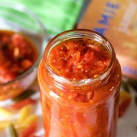 A jar of orangey red tomato basil pasta sauce with a bowl of sauce behind it and a cook book to one side. Coloured penne pasta scattered all around and a green edged napkin at the back