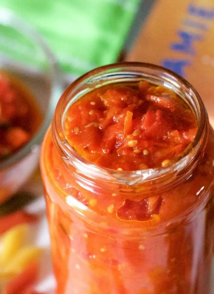 A jar of orangey red tomato basil pasta sauce with a bowl of sauce behind it and a cook book to one side. Coloured penne pasta scattered all around and a green edged napkin at the back