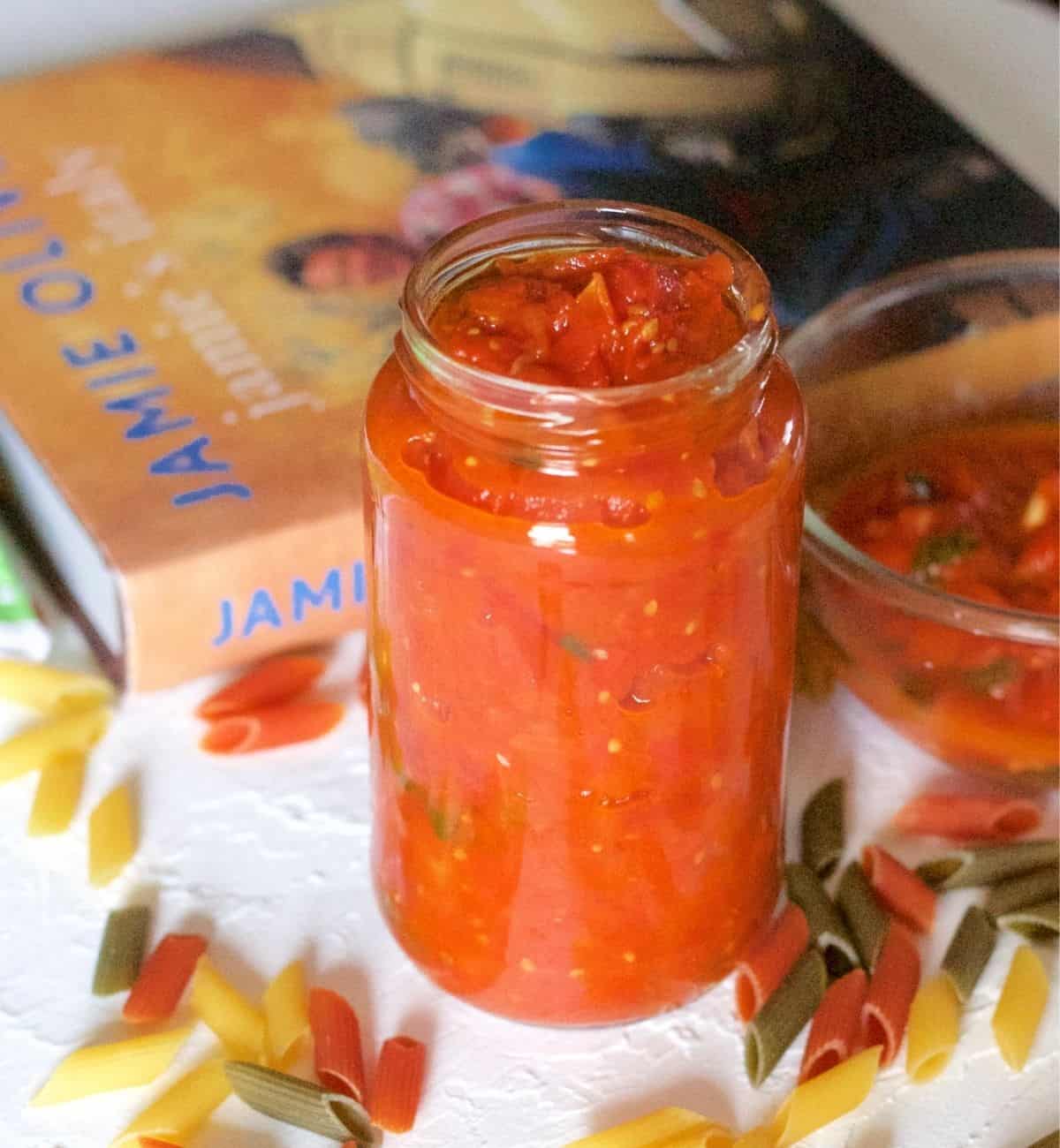 Orange red tomato basil pasta sauce in a tall glass jar with a yellow/orange covered cook book in the background. A bowl of sauce on the right and coloured penne pasta scattereda all around