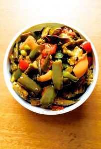 Bright coloured brinjal curry with capsicum and red and yellow bell peppers and grean peas ina white bowl against a pale orange background