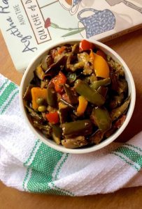 A white bowl of brinjal curry with green, red and yellow peppers and green peas, on a pale orange backrground. A white napkin with broad green stripes is placed just below the bowl and to the left, and part of a book can be seen on the top right with the words Agatha and Quiche partly visible