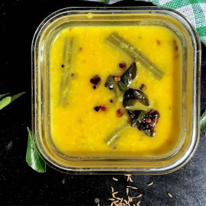 Bright yellow easy vegetable Kootu for the new Mom in a square glass bowl. Slices of green drumstick on the surface of the lentils, and a dark brown tempering of cumin, mustard, fenugreek and curry leaves on top