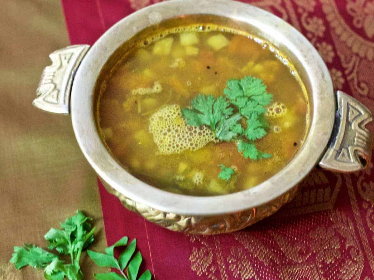 PIneapple rasam, a pale mustardy yellow coloured soup like liquid in a brass bowl,with coriander leaves as garnish. Resting on a red and olive green silk fabric