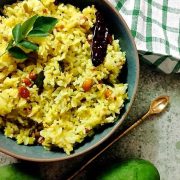 Yellow Raw mango rice or mangai sadam from grated seasonal raw green mango, seasoned with mustard, peanuts, dry chili and curryleaves, in a green bowl wiht an arrow shaped brass spoon on one side, a green mango and white and green checked napkin above