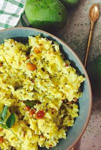 Bright yellow raw mango rice seasoned with penauts, curry leaves, lentils,mustard seeds, in a green bowl. A brass spoon seen to the right and part of a checked green on white napkin to the left