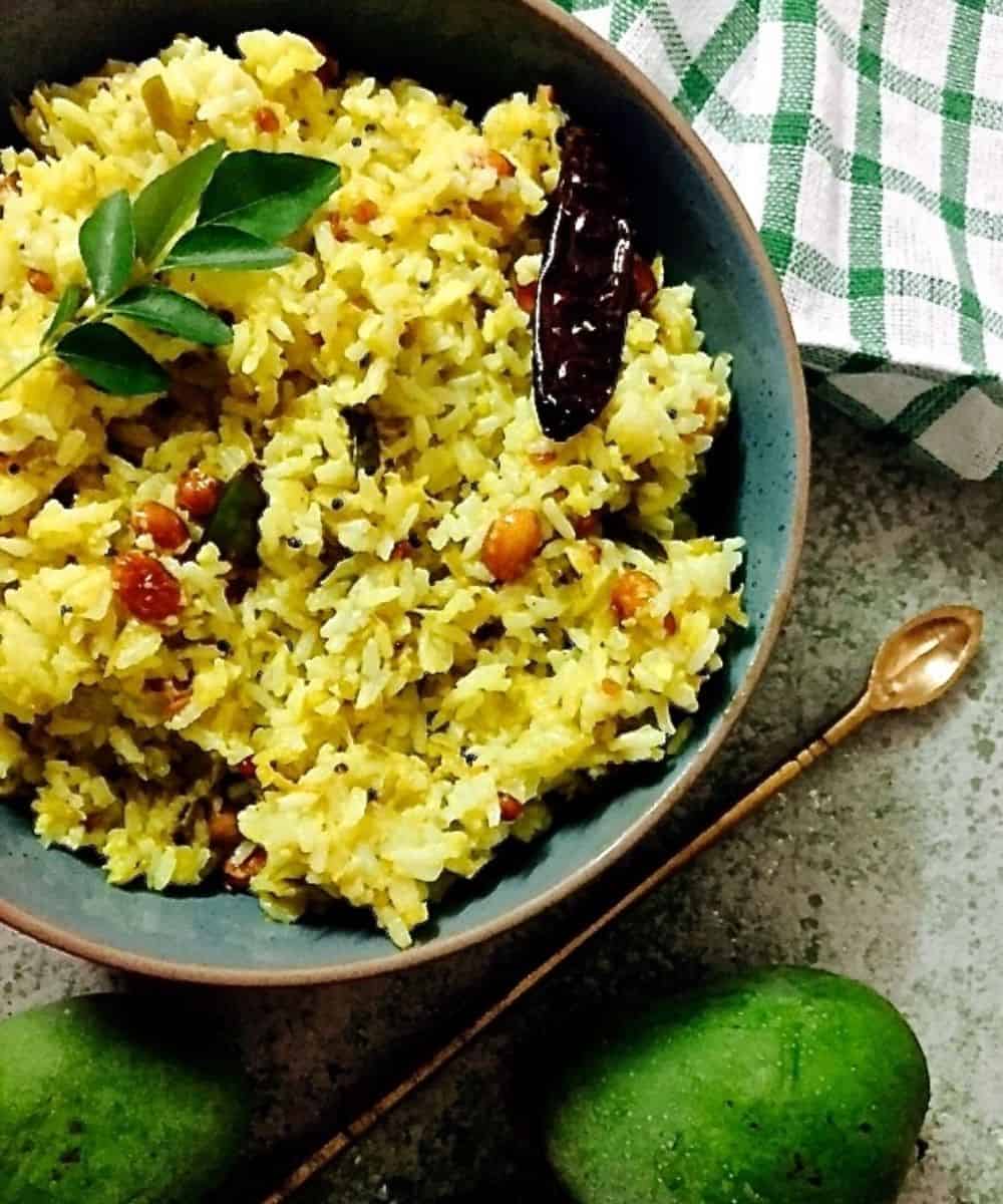 Green blue Bowl of yellow mango rice garnished with a sprig of curry leaves and with fried peanuts. A green and white checked napkin in the background, 2 green mangoes in the foreground and a brass narrowheaded spoon on the right.