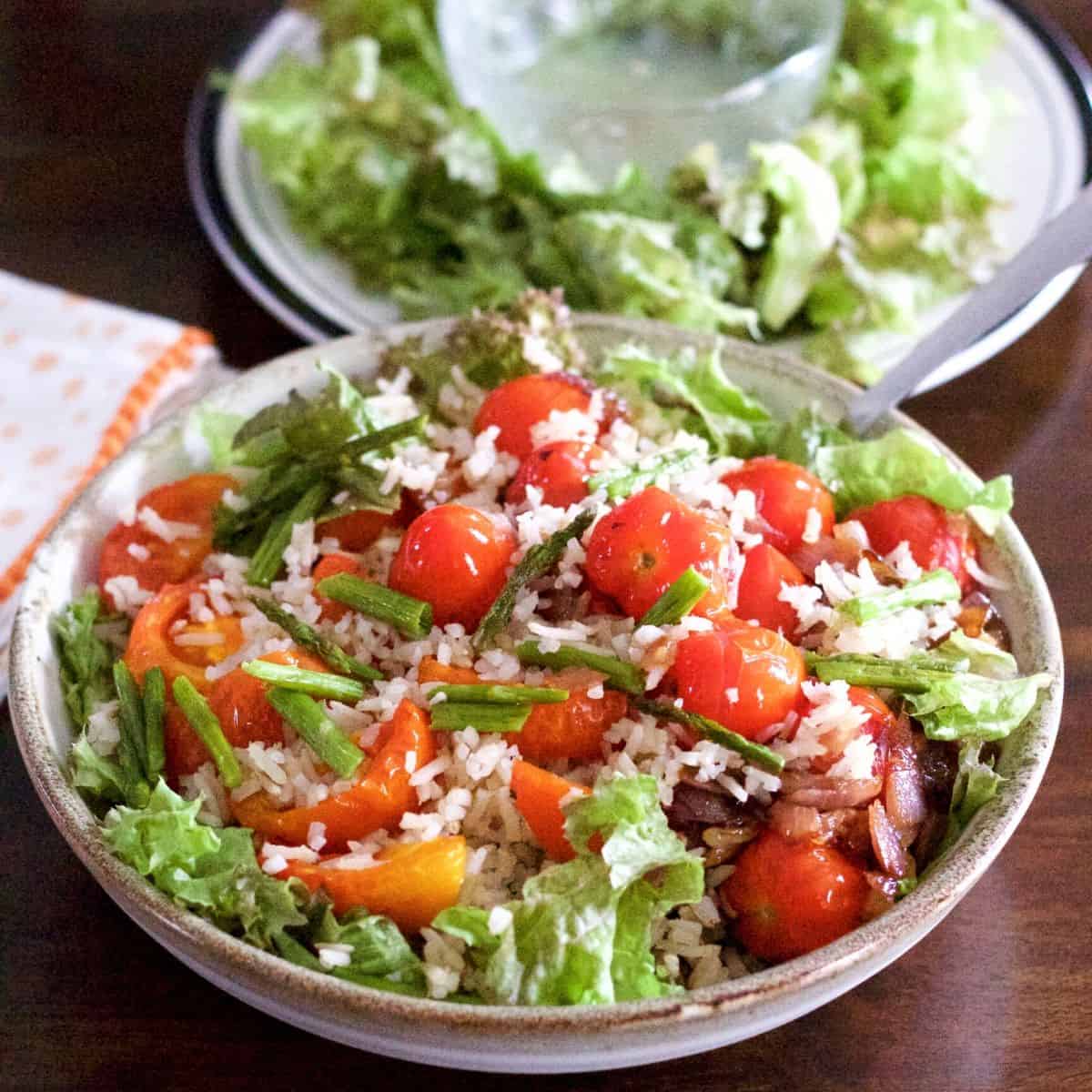 Bowl filled with red cherry tomatoes piled on a salad of brown rice, roasted tomato slices and greens. With a plate of greens and a glass bowl of lemon dressing in the background. Par of an orange edged white napkin on the left. All on a dark brown background