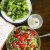 A bowl of red cherry tomatoes heaped on a salad of brown rice and salad greens. A small bowl of lemon dressing with a red chili and a blue bordered plate with greens, in the background. A white napkin with orange dots on the left. https://www.PepperOnPizza.com