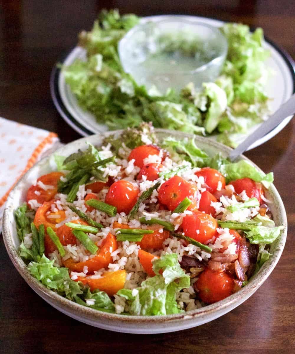 Red cherry tomatoes and salad greens with brown rice heaped on a white bowl. A blue rimmed plate of greens and a glass bowl with lemon dressing in the background. An orange dotted white napkin to the left