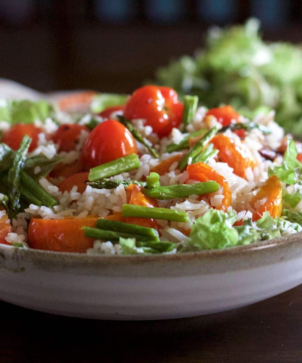 Large bowl of salad of tomato, brown rice, salad greens heaped on it. A blue rimmed plate of salad greens and a glass bowl with lemon dressing at the back and a orange dotted white napkin to the left