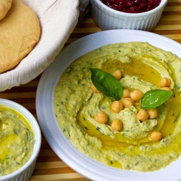 Fresh Lemon Basil Hummus with garnish of olive oil and basil leaves on a white plate with pita bread and cherry dip