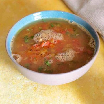 A bowl of orange red liquid, rasam with pineapples and tomato and coriander leaves in a blue edged bowl on a yellow background. A beige napkin in the top right