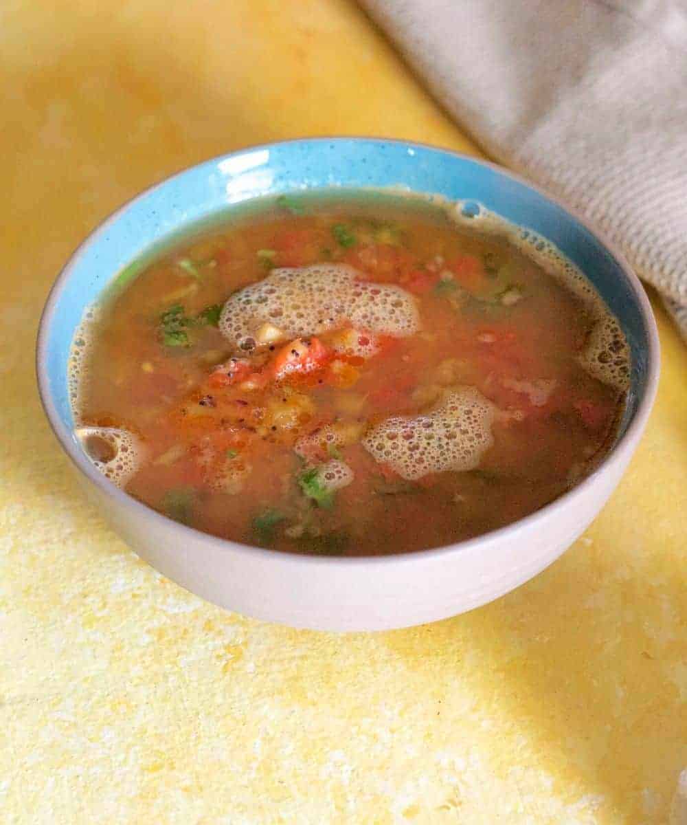 A bowl of orange red liquid, rasam with pineapples and tomato and coriander leaves in a blue edged bowl on a yellow background. A beige napkin in the top right