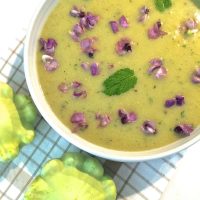 A white edged bowl of green yellow patty pan soup, garnsihed with fresh purple hyacinth flowers and fresh mint leaves. Two green patty pan squash seen on the thin brown striped white napkin in the foreground