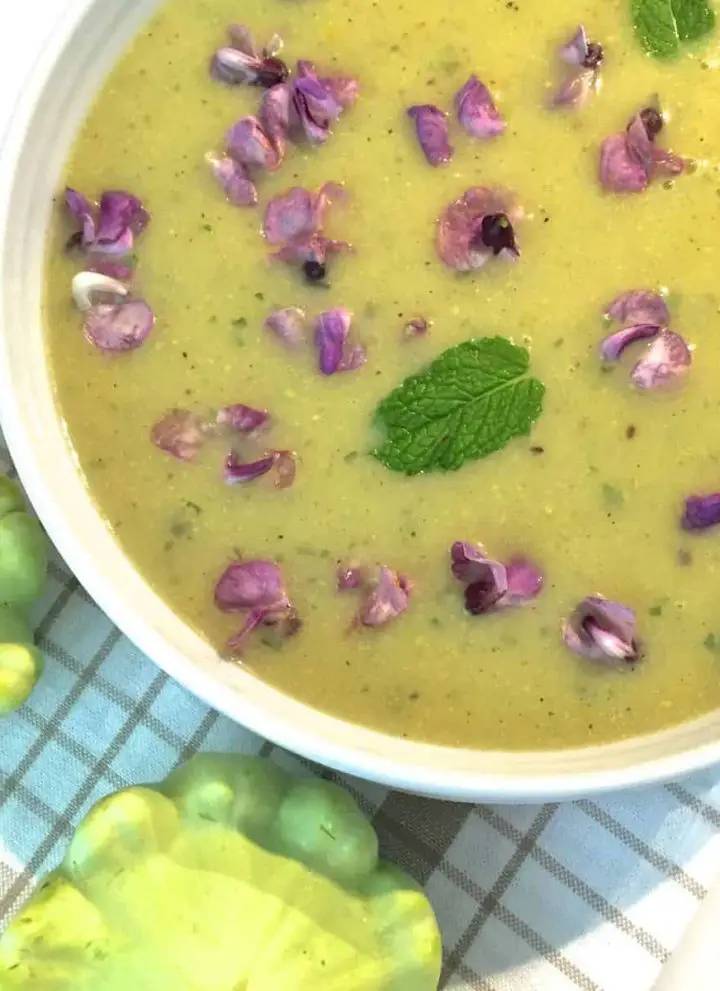 A white edged bowl of green yellow patty pan soup, garnsihed with fresh purple hyacinth flowers and fresh mint leaves. Two green patty pan squash seen on the thin brown striped white napkin in the foreground