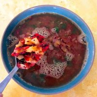 Reddish brown soup like pineapple and tomato rasam in a blue bowl with a spoon showing tempering of pineapple and tomato. All on a yellow background