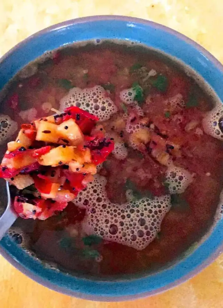 Reddish brown soup like pineapple and tomato rasam in a blue bowl with a spoon showing tempering of pineapple and tomato. All on a yellow background