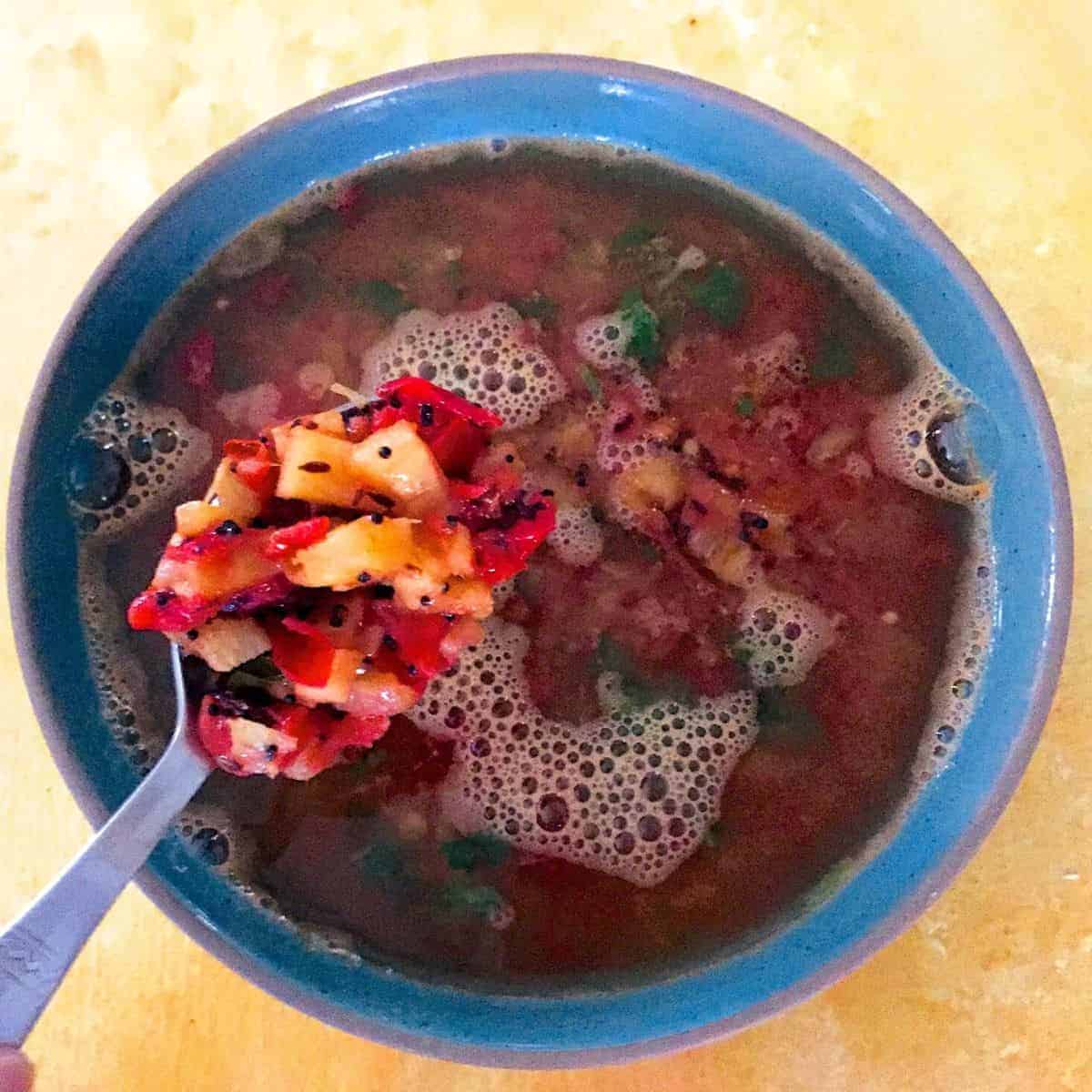 Reddish brown soup like rasam in a blue bowl with a spoon showing tempering of pineapple and tomato. All on a yellow background