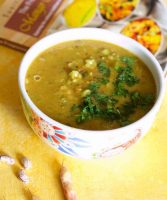 A large white bowl with red and blue pattern on the side, with mustard coloured lentil stew, nourishing masoor dal for the new mom.A cookbook with orange and yellow design in the background, a few pink cranberry beans in the foreground, the whole on a yellow background