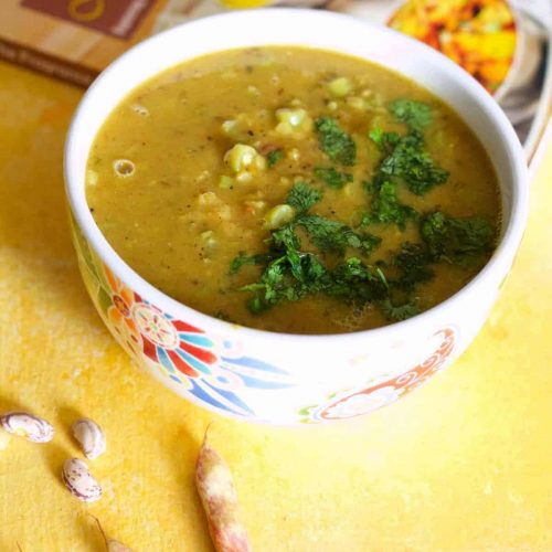 Mustard coloured lentil stew, Nourishing Masoor dal for the New Mom, in a white bowl patterned with red and blue, on a yellow background. Pink cranberry beans in the foreground and a red and orange covered cook book in the background