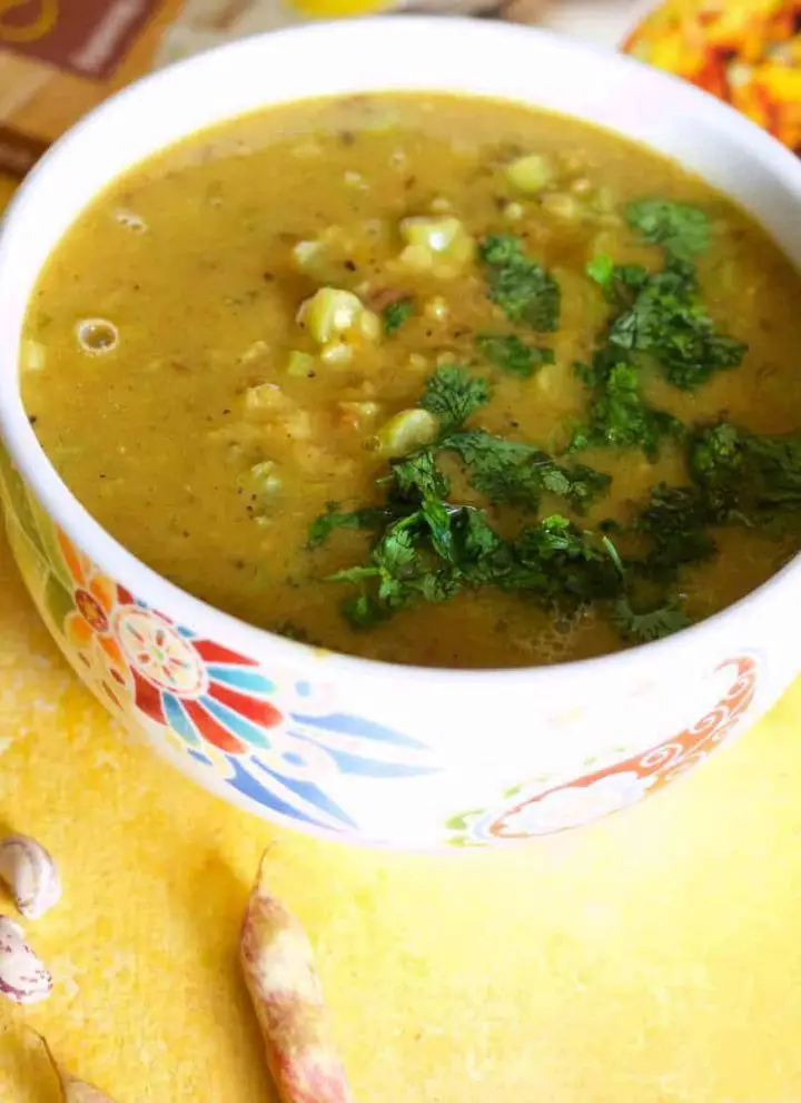 Mustard coloured lentil stew, Nourishing Masoor dal for the New Mom, in a white bowl patterned with red and blue, on a yellow background. Pink cranberry beans in the foreground and a red and orange covered cook book in the background