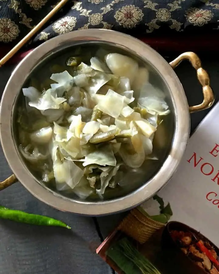 Oying Vegetable Stew from Arunachal Pradesh in NE India. Brass bowl with stew of cabbage, potato and beans with Cookbook and a green chilli by the side