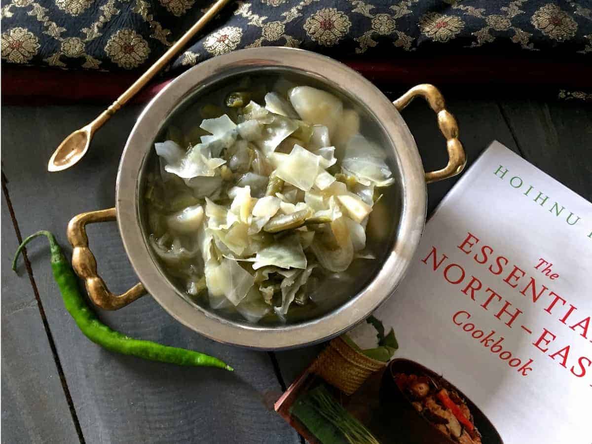 Oying Vegetable Stew from Arunachal Pradesh in NE India. Brass bowl with stew of cabbage, potato and beans with Cookbook and a green chilli by the side