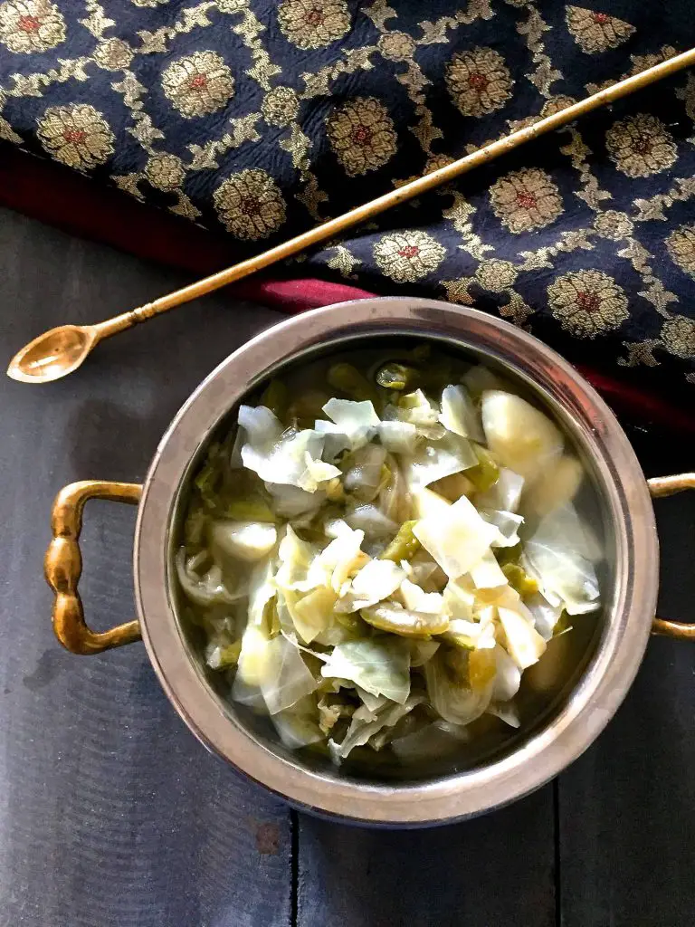 Brass bowl with handles with potato cabbage beans stew and a black and red silk cloth above with an arrow shaped spatula lying across