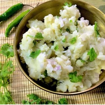 Aloo Pitika_F Assamese Aloo Pitika - a brass bowl with mashed potatoes, cilantro, mustard oil, green chillies and onion on a mat, with green chillies and cilantro alongside