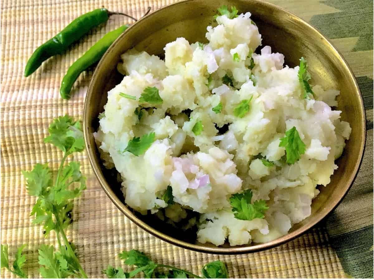 Assamese Aloo Pitika - a brass bowl with mashed potatoes, cilantro, mustard oil, green chillies and onion on a mat, with green chillies and cilantro alongside