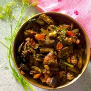 Baingan Badi Sabzi - curried slices of aubergine with black gram lentil fritters, turmeric, spinach, dill and Indian spices, in a brass bowl with dill leaves alongside and resting near a pink embroidered cloth