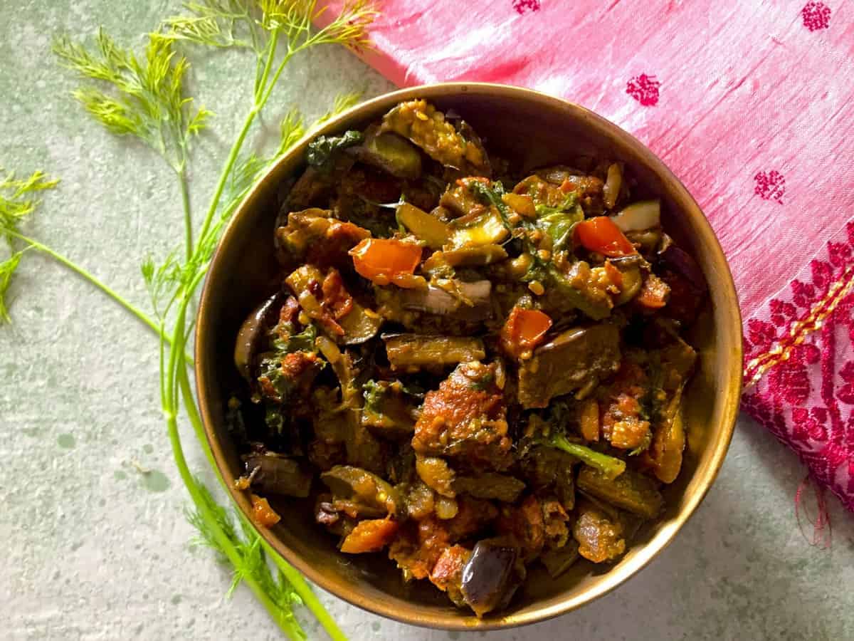 Baingan Badi Sabzi - curried slices of aubergine with black gram lentil fritters, turmeric, spinach, dill and Indian spices, in a brass bowl with dill leaves alongside and resting near a pink embroidered cloth