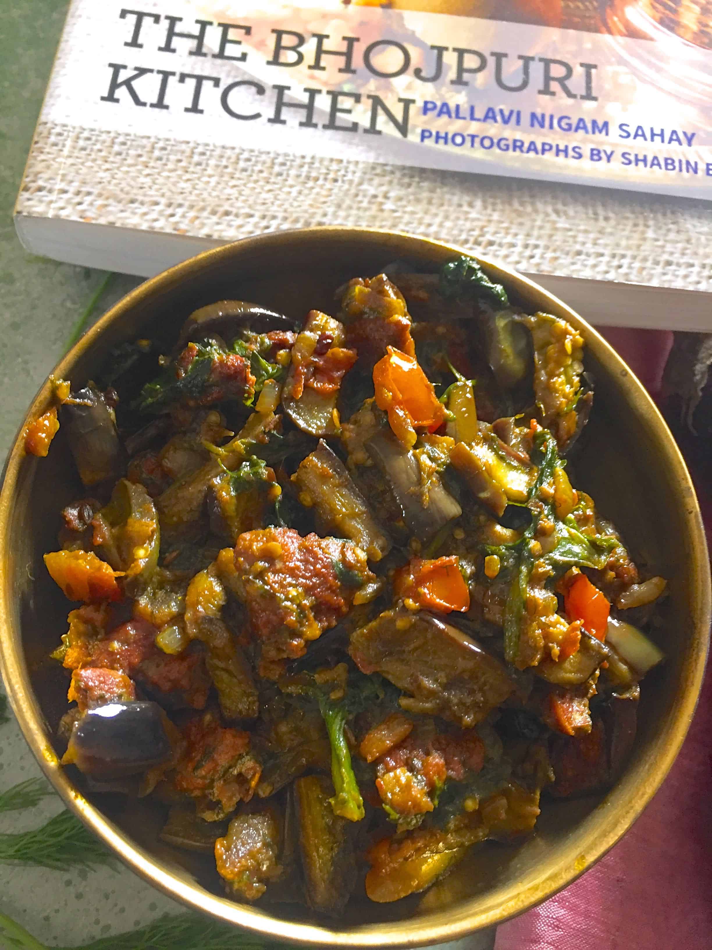 Brass bowl filled with brinjal curry with red tomatoes, lentil fritters and dill. A book on Bihari Cuisine in the background, a pale pink emroidered saree in the background