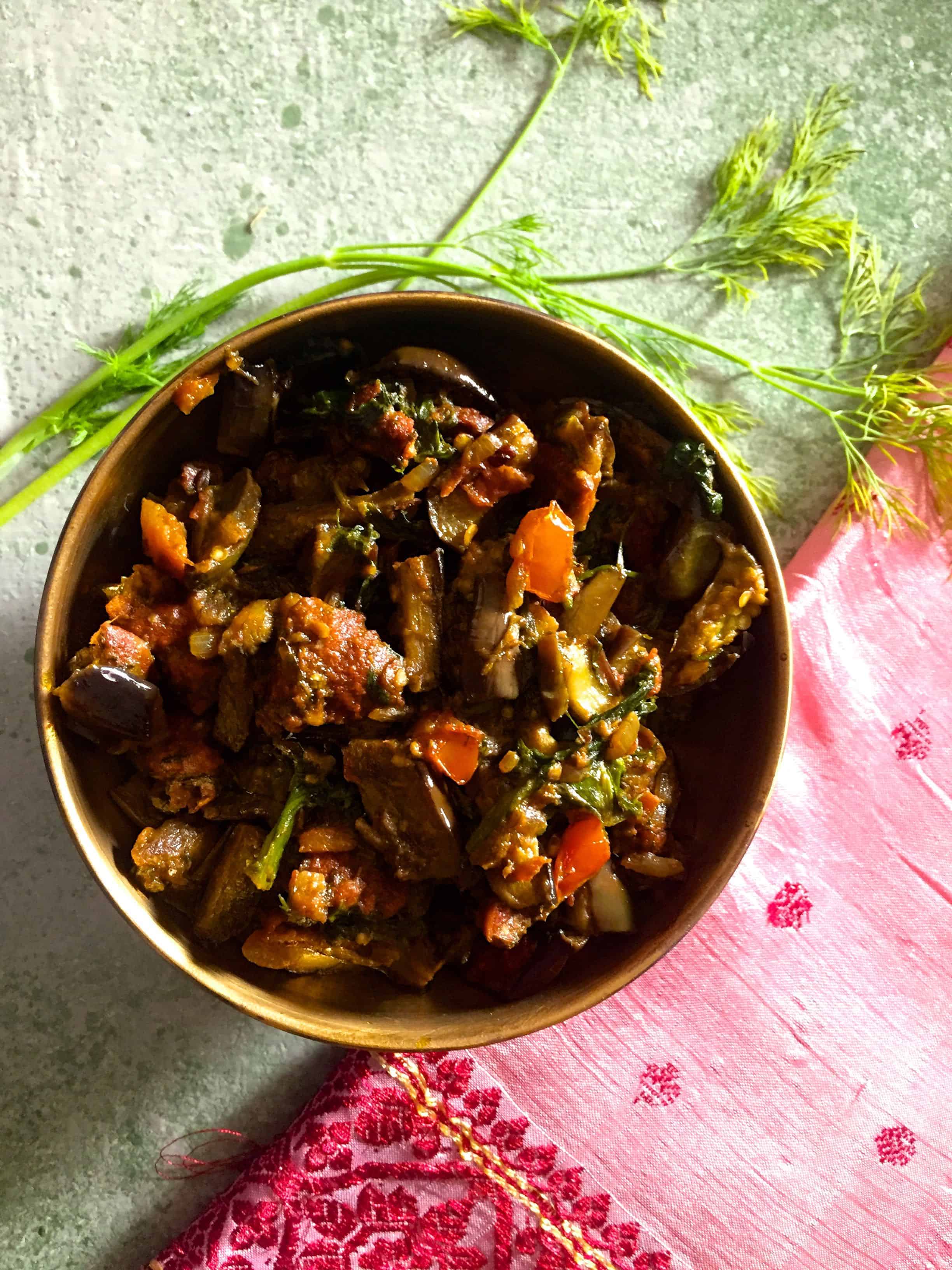 A bowl of baingan badi curry on a green background with dill leaves on one side and a pink embroidered fabric alongside