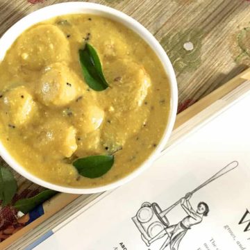 A bowl of creamy yellow colocasia in yogurt or arbi ki kadhi from the cuisine Indian State Chattisgarh State. The white bowl rests on a brown mirror worked fabric with a book by the side with a sketch of a person churning butter