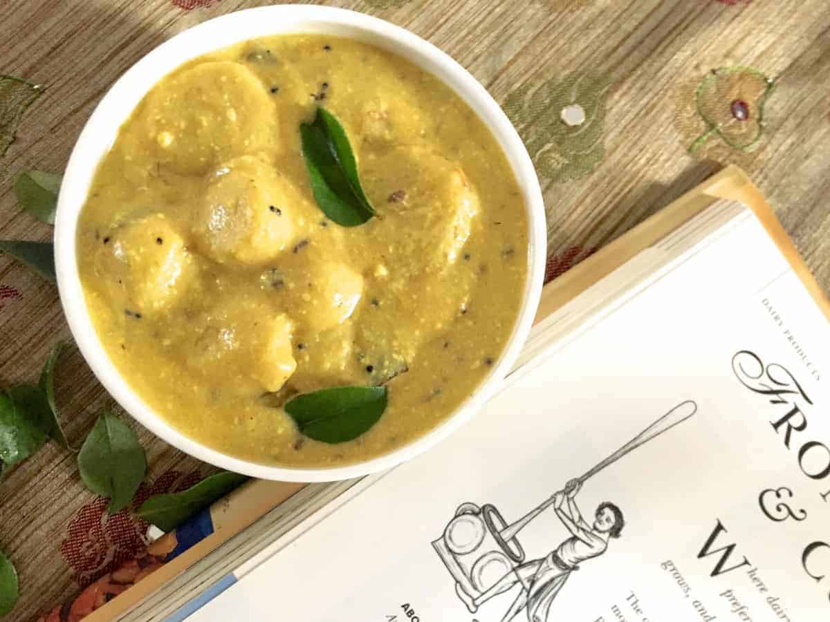 A bowl of creamy yellow colocasia in yogurt or arbi ki kadhi from the cuisine Indian State Chattisgarh State. The white bowl rests on a brown mirror worked fabric with a book by the side with a sketch of a person churning butter