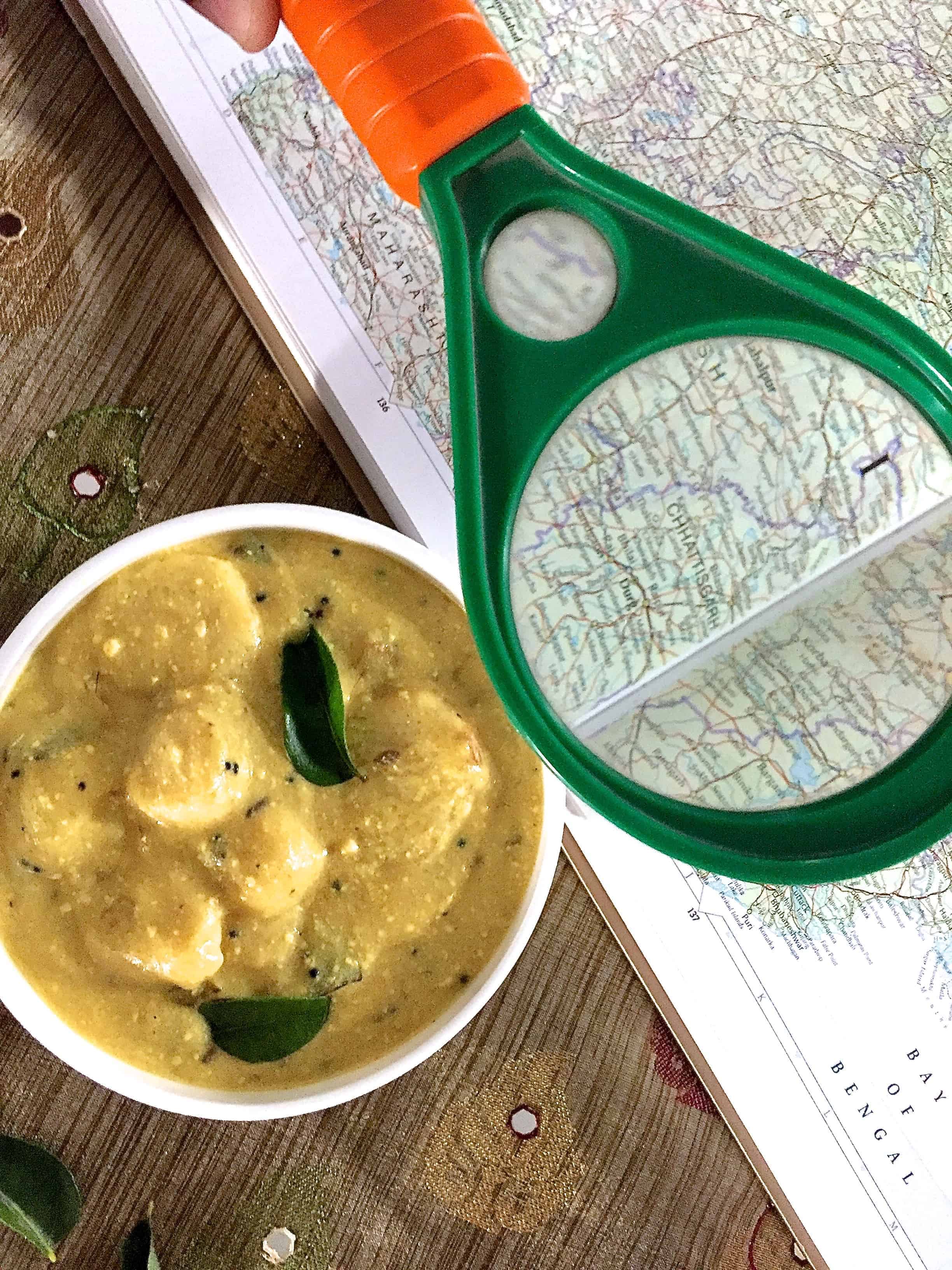 White bowl filled with arbi ki kadhi, a yellow curd based gravy with colocasia root/ arbi, mustard seeds and curry leaves seen on top. An open atlas with a magnifying glass highlights Indian State Chattisgarh